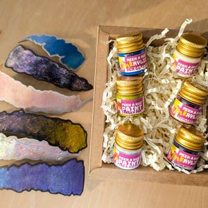 overhead view of a cheerful kraft gift box on a wood surface filled w/ 6 petite glass jars of sparkly iridescent paint. Each jar has a different color and a colorful label on it, and a matte metallic twist off gold lid. The box is filled with recycled off-white crinkle paper and flanked by shimmering color-shifting shaped swatches of each paint color on the left of the image.