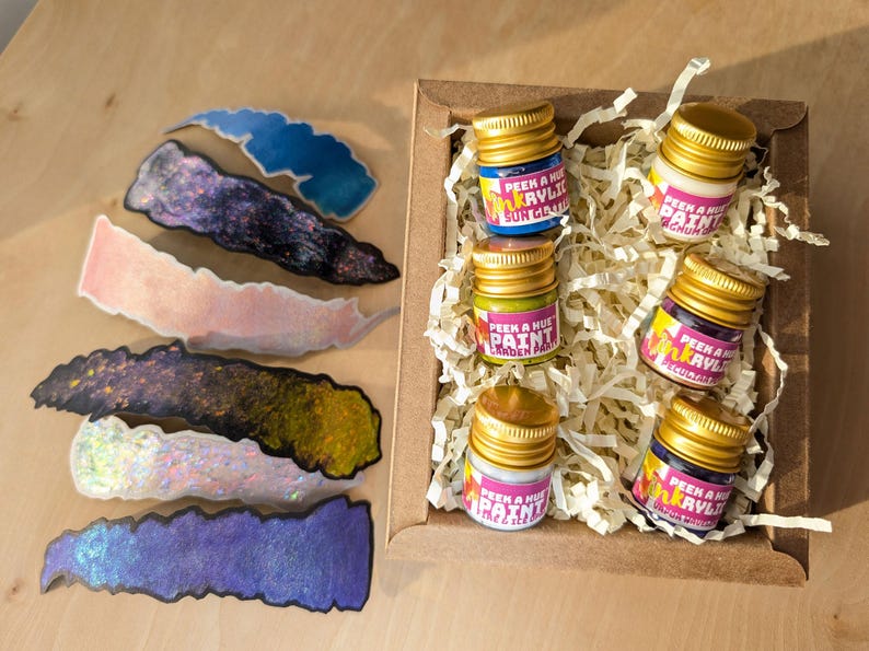 overhead view of a cheerful kraft gift box on a wood surface filled w/ 6 petite glass jars of sparkly iridescent paint. Each jar has a different color and a colorful label on it, and a matte metallic twist off gold lid. The box is filled with recycled off-white crinkle paper and flanked by shimmering color-shifting shaped swatches of each paint color on the left of the image.
