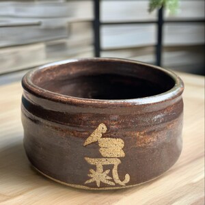 May include: A round, dark brown ceramic bowl with a glossy, mottled finish. The exterior features a prominent, textured gold-colored Japanese kanji character for 'ki' or energy, applied in a raised, matte style against the smooth, dark clay surface.