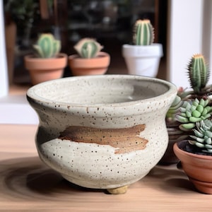 May include: A round, speckled off-white ceramic planter with a wide, flared rim and a single horizontal brown brushstroke accent across the front. The planter features a rounded body and is supported by small, short feet at the base.