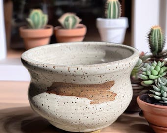 Peut inclure: Un pot de fleurs en céramique blanc cassé moucheté, doté d'un bord large et évasé ainsi que d'un coup de pinceau brun horizontal sur le devant. Le pot présente un corps arrondi et repose sur de petits pieds courts à la base.