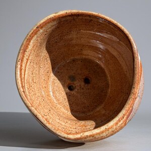 Peut inclure: Un bol en c&eacute;ramique fait &agrave; la main, mouchet&eacute;, avec un large bord et deux trous de drainage. Le bol pr&eacute;sente une palette de couleurs chaudes et terreuses avec des nuances de brun et d'orange. L'int&eacute;rieur a une finition lisse, tandis que l'ext&eacute;rieur a un aspect textur&eacute;.