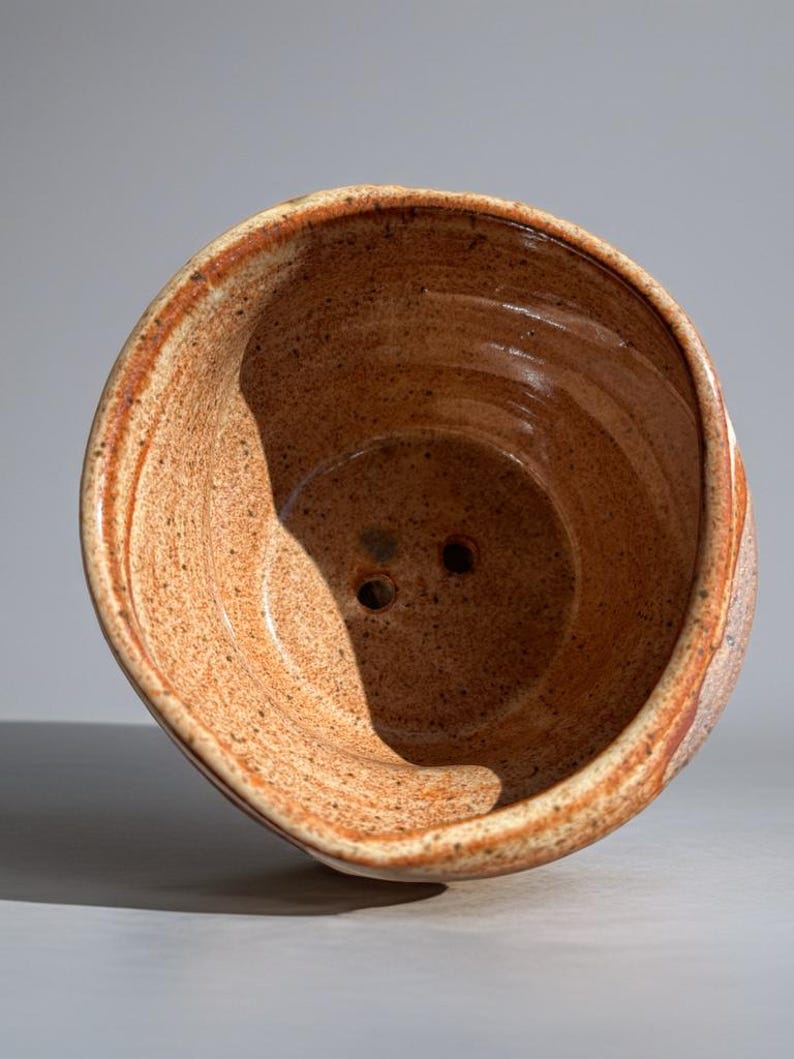 Peut inclure: Un bol en c&eacute;ramique fait &agrave; la main, mouchet&eacute;, avec un large bord et deux trous de drainage. Le bol pr&eacute;sente une palette de couleurs chaudes et terreuses avec des nuances de brun et d'orange. L'int&eacute;rieur a une finition lisse, tandis que l'ext&eacute;rieur a un aspect textur&eacute;.