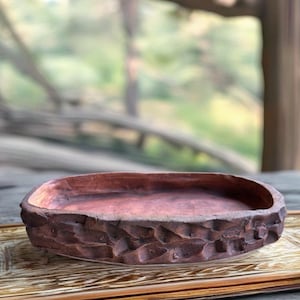 May include: A handcrafted, oval-shaped, brown wooden bowl with a textured, carved exterior. The bowl sits on a wooden tray, showcasing a natural, rustic aesthetic. The interior of the bowl has a smooth, reddish-brown finish.