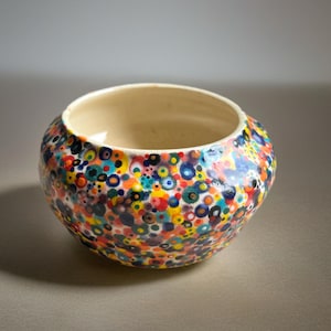 May include: A round ceramic bowl with a speckled pattern of colorful dots in shades of blue, yellow, red, and green. The interior of the bowl is a light beige color.