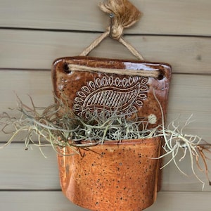 May include: A hanging ceramic planter with a brown, speckled finish. The planter features a decorative paisley design and is filled with dried plants. It is suspended by a natural rope, perfect for adding a rustic touch to any space.