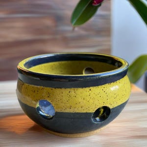 May include: A round ceramic bowl featuring horizontal stripes of speckled yellow and dark gray glaze. The bowl has several circular cutouts along its sides, revealing the interior space and the smooth, rounded rim at the top.
