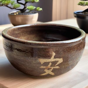 May include: A round, brown ceramic bowl with a glossy, mottled glaze finish. The exterior features a single, large, light-colored Japanese kanji character, 安, centered on the front. The bowl has a thick, rounded rim and a deep, dark interior.