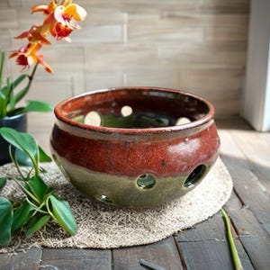 Op de afbeelding: Een handgemaakte keramische kom met een tweekleurig glazuur. Het bovenste gedeelte is een diep roodbruin, overgaand in een groene basis. De kom heeft verschillende cirkelvormige openingen. Een orchideeplant met oranje en gele bloemen staat op de achtergrond.