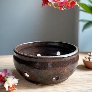 May include: A dark brown ceramic bowl with a glossy finish and multiple circular drainage holes along the lower sides. The bowl has a rounded shape with a thick rim and a smooth, reflective surface that catches the light, showing subtle color variations.