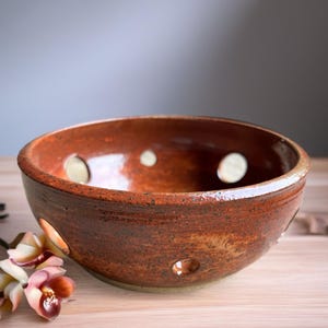 May include: A round, glazed ceramic bowl with a deep reddish-brown, speckled finish. The bowl features several circular cutouts of varying sizes distributed around its sides, creating a perforated appearance with a smooth, reflective surface.
