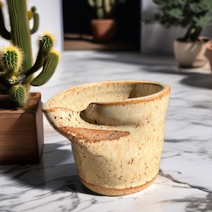 May include: A cream-colored ceramic creamer with a speckled finish and a distinct, wide pouring spout. The vessel features a textured, earthy brown rim and base, with a hand-formed, asymmetrical shape that tapers slightly toward the bottom.