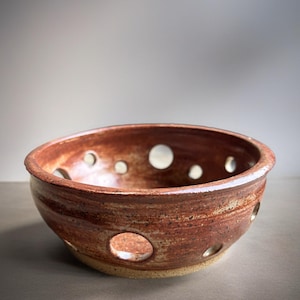 May include: A round, hand-thrown ceramic bowl with a glossy, variegated reddish-brown glaze. The bowl features a series of circular cutouts spaced evenly around its sides, with a smooth, rounded rim and a natural, unglazed tan base.