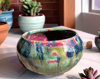 Peut inclure: Un bol en céramique rond présentant un émail brillant et multicolore avec des traînées mélangées de vert vibrant, de bleu profond et de rose vif. L'intérieur du bol est sombre et la surface extérieure a une finition lisse et réfléchissante.