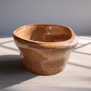 May include: A handmade ceramic bowl with a unique, organic shape. The bowl features a warm, earthy color palette with shades of brown and tan, and a textured surface. The rim has a lighter, almost white, accent. The bowl is suitable for serving or display.