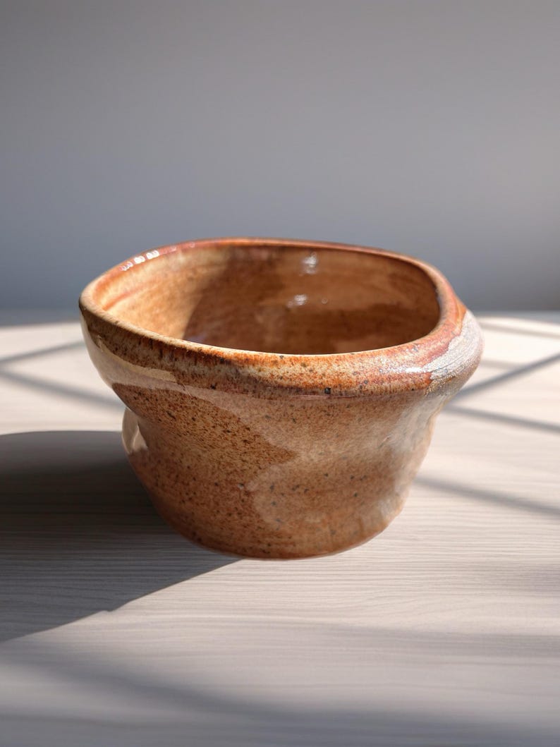 Peut inclure: Un bol en c&eacute;ramique fait &agrave; la main avec une forme unique et organique. Le bol pr&eacute;sente une palette de couleurs chaudes et terreuses avec des nuances de brun et de beige, et une surface textur&eacute;e. Le bord a un accent plus clair, presque blanc. Id&eacute;al pour servir ou exposer.
