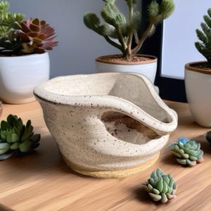 May include: A handmade ceramic yarn bowl with a speckled cream glaze and a smooth, unglazed tan base. The bowl features a deep, curved cutout on one side designed for guiding yarn, with a wide, rounded rim and a sturdy, thick-walled construction.