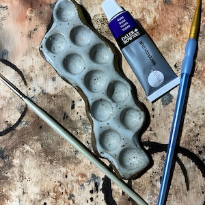 May include: A ceramic paint palette with multiple wells, a tube of Daler Rowney Simply Watercolour in Violet, and two paint brushes. The palette is light blue with speckled detailing. One brush has a blue handle and the other has a light green handle.