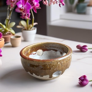 May include: A round ceramic bowl with a speckled white interior and a glossy, mottled brown and gold exterior glaze. The bowl features several circular cutouts along its sides, creating a distinct, open-work design with a thick, textured rim.