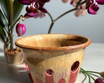 Peut inclure: Un pot de fleurs en céramique rond avec un bord large, doté d'un vernis brillant passant d'un jaune miel chaud en haut à un brun rougeâtre profond à la base. Le pot présente plusieurs trous de drainage circulaires sur les côtés.