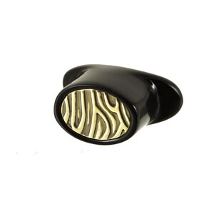 May include: An oval-shaped cabinet knob with a glossy black finish and a central gold-toned inlay featuring a raised, textured, vertical wavy line pattern. The knob has a smooth, rounded outer edge and a wide, flat base for mounting.