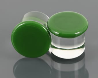 Puede incluir: Dos tapones para orejas de vidrio verde con bases de vidrio transparente. Los tapones son redondos y tienen un acabado liso y pulido.