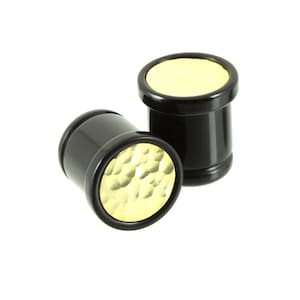 May include: Two black ear plugs with gold-coloured, textured faces. The plugs are cylindrical with a slightly flared edge. One plug is lying on its side, while the other stands upright, both against a white background.
