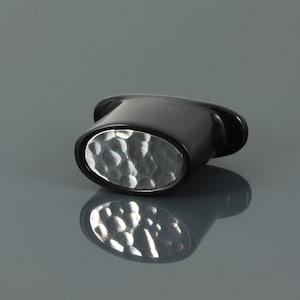 Puede incluir: Un tapón de botella ovalado negro con un inserto de metal martillado plateado.