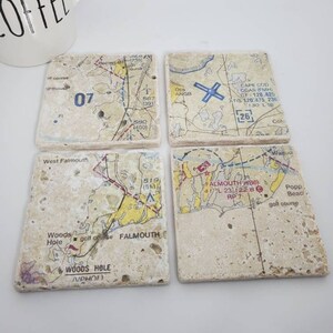 May include: Set of four coasters featuring vintage maps of Falmouth, Massachusetts. Each coaster is made of beige travertine stone with a printed map design. The maps show various landmarks, including Woods Hole, West Falmouth, and the Otis ANGB.