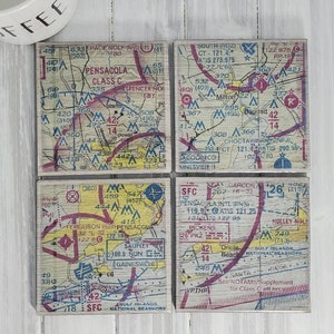 May include: Four square coasters with vintage-style maps of Pensacola, Florida. Each coaster features a different section of the city, including the airport, the Gulf Islands National Seashore, and the surrounding area. The maps are printed in red, blue, yellow, and black on a wood-grain background.