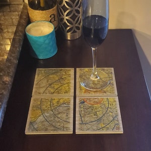 May include: Four square coasters with a vintage map design. The coasters are made of wood and have a light brown color. The map design features lines, numbers, and text. A glass of red wine sits on top of the coasters.