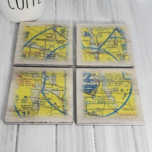 May include: Four square coasters with a vintage map design. Each coaster features a different section of a map with blue, yellow, and white markings. The coasters are made of wood with a whitewashed finish.