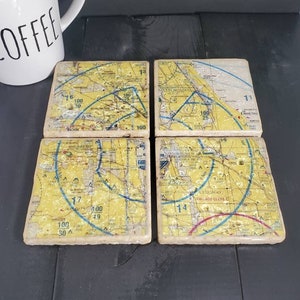May include: Set of four square coasters featuring vintage-style maps with yellow, blue, red, and white markings. The maps include numbers, text, and lines. The coasters are made of a light brown material.
