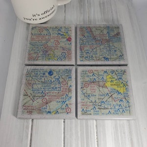 May include: Four square coasters with a vintage map design. Each coaster features a different section of a map with various locations, including Dothan, Alabama, and surrounding areas. The maps are printed in black, blue, red, and white on a light brown background.