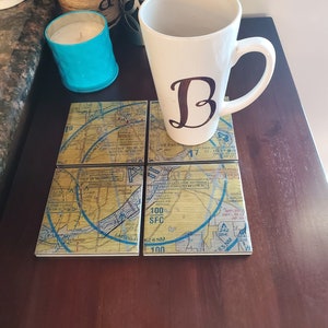 May include: Four square coasters with a map design featuring blue and yellow colors. The coasters are made of ceramic and have a glossy finish. The coasters are arranged in a square shape with a white mug with a black letter "B" on top of the coasters.