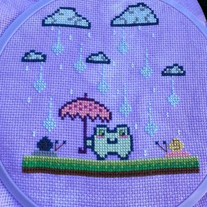 May include: Cross-stitch embroidery featuring a frog sheltering under an umbrella in the rain. The frog is green and white, and the umbrella is pink. The background is a light purple fabric, framed in a purple hoop.