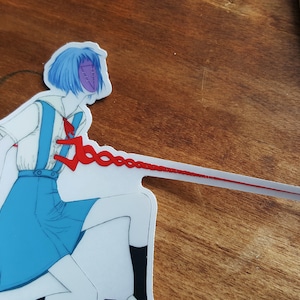 May include: A white sticker with a cartoon illustration of a girl with blue hair wearing a blue and white school uniform. The girl has a purple mask with a stitched design. A red string is attached to the girl's chest and extends to the right side of the sticker.