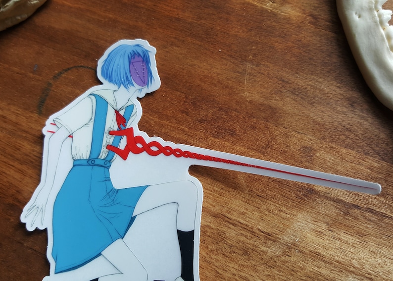 May include: A white sticker with a cartoon illustration of a girl with blue hair wearing a blue and white school uniform. The girl has a purple mask with a stitched design. A red string is attached to the girl's chest and extends to the right side of the sticker.