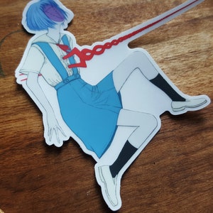 May include: A sticker featuring a blue-haired anime character in a white and blue school uniform, with a red chain attached to their chest. The character is drawn in a dynamic pose, with one leg raised and their arms outstretched.
