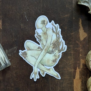 May include: A sticker featuring a detailed illustration of a pale, human-like figure with a face obscured by a cluster of small, round shapes. The figure is depicted in a seated position with its arms and legs covered in intricate, organic patterns resembling coral or seaweed.