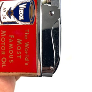 Op de afbeelding: Een rode en gouden aansteker met een zilveren bovenkant. De aansteker heeft een afbeelding van een blik Veedol motorolie met de tekst "The World's Most Famous Motor Oil" op de voorkant.
