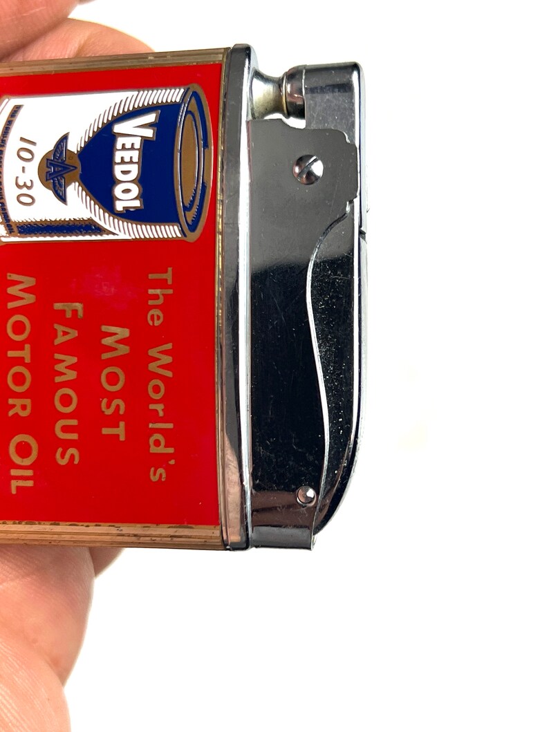 Op de afbeelding: Een rode en gouden aansteker met een zilveren bovenkant. De aansteker heeft een afbeelding van een blik Veedol motorolie met de tekst "The World's Most Famous Motor Oil" op de voorkant.