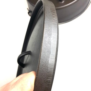 Puede incluir: Primer plano de una tapa de hierro fundido negra con una superficie texturizada y un asa. La tapa est&aacute; siendo sostenida por una mano.