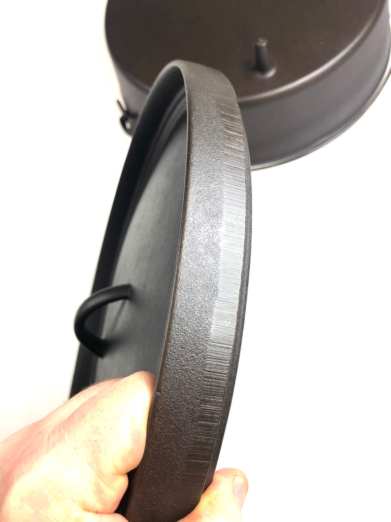 Puede incluir: Primer plano de una tapa de hierro fundido negra con una superficie texturizada y un asa. La tapa est&aacute; siendo sostenida por una mano.