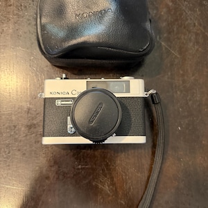 May include: A vintage Konica C35 camera with a black leather case and lens cap. The camera body is silver and black, with the Konica logo visible on the lens cap and case. A black wrist strap is attached to the camera.