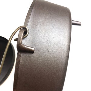 Puede incluir: Una olla de hierro fundido de color gris oscuro con tapa y asa. La olla tiene tres patas y un bucle en el lateral para colgarla.