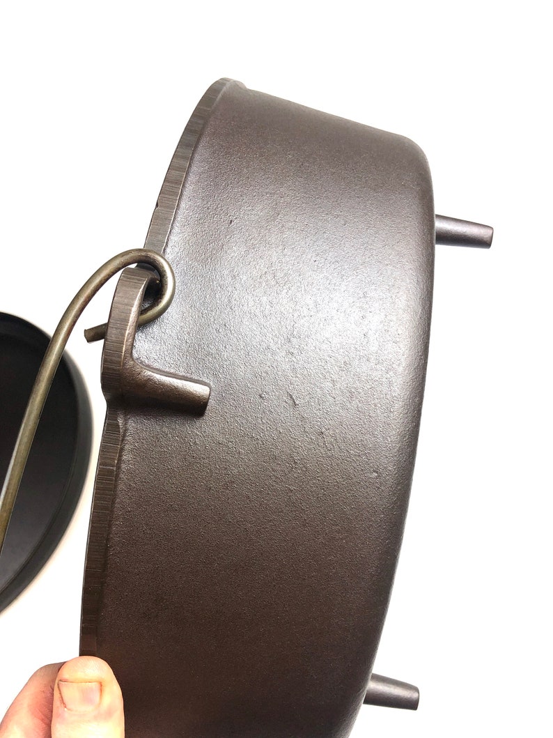 Puede incluir: Una olla de hierro fundido de color gris oscuro con tapa y asa. La olla tiene tres patas y un bucle en el lateral para colgarla.