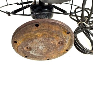 May include: An antique electric fan with a black motor housing, blades, and protective cage. The base is a rusty brown color, and the fan has a black power cord. The fan blades are visible within the cage.