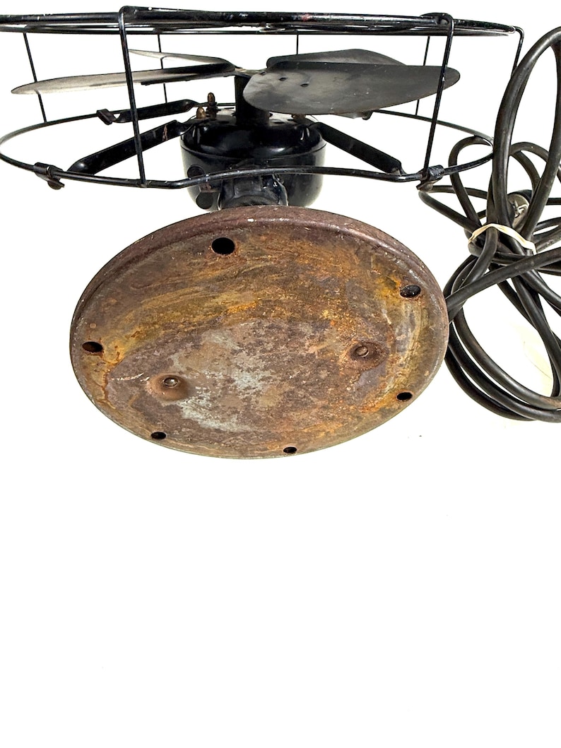May include: An antique electric fan with a black motor housing, blades, and protective cage. The base is a rusty brown color, and the fan has a black power cord. The fan blades are visible within the cage.