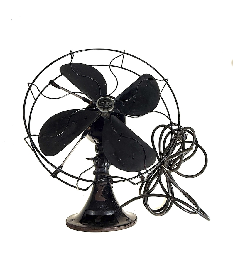 May include: A vintage black Emerson electric fan. The fan has four blades and a protective wire cage. The base is round and the fan is connected to a black power cord. The fan is a classic design.